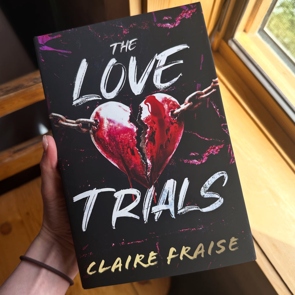 A hand holds a book titled 'The Love Trials' by Claire Fraise. The book cover is black with a cracked red heart and white title text, photographed in a warm indoor setting near a window.