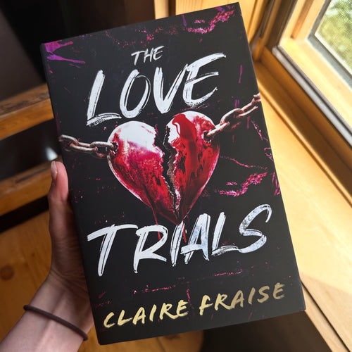 A hand holds a book titled 'The Love Trials' by Claire Fraise. The book cover is black with a cracked red heart and white title text, photographed in a warm indoor setting near a window.